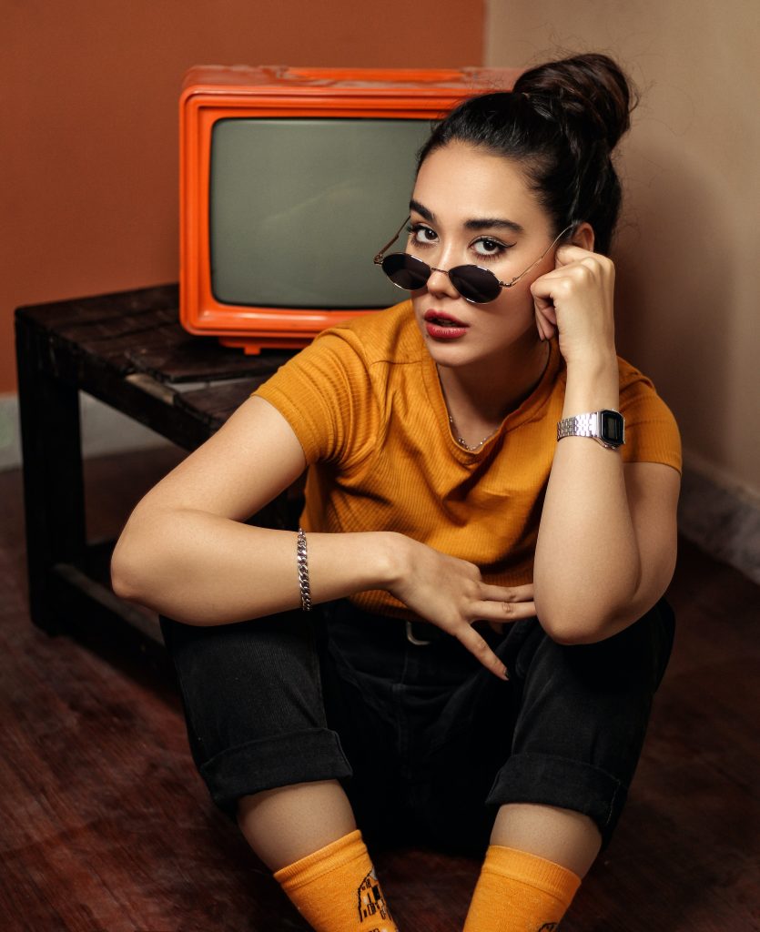 Fată cu ochelari de soare în tricou portocaliu, așezată pe jos, lângă un televizor.