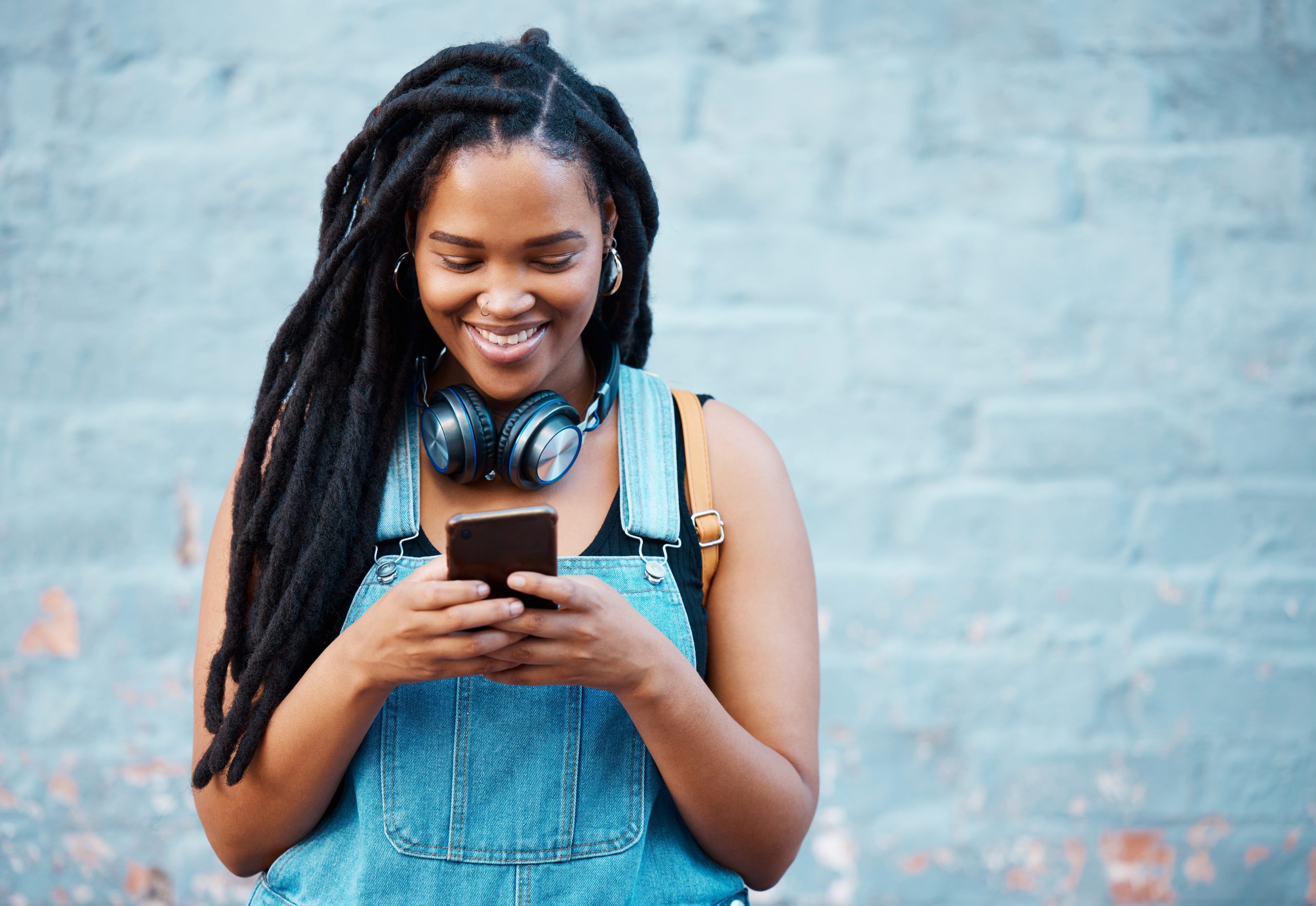 Fată afro-americană, îmbrăcată în salopetă albastră care ascultă Taylor Swift la căști și se uită pe TikTok pe telefon și zâmbește.